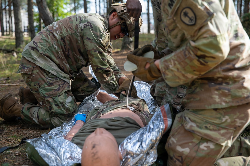 Two U.S. Army soldiers in OCP (Operational Camouflage Pattern) uniforms are providing tactical combat casualty care (TCCC) to a downed soldier lying on a mylar/emergency thermal blanket on the ground. The medics appear to be conducting a field medical training exercise or responding to a simulated casualty in a wooded outdoor environment. One medic is wearing blue medical gloves and both are actively treating the patient.