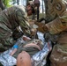 Two U.S. Army soldiers in OCP (Operational Camouflage Pattern) uniforms are providing tactical combat casualty care (TCCC) to a downed soldier lying on a mylar/emergency thermal blanket on the ground. The medics appear to be conducting a field medical training exercise or responding to a simulated casualty in a wooded outdoor environment. One medic is wearing blue medical gloves and both are actively treating the patient.