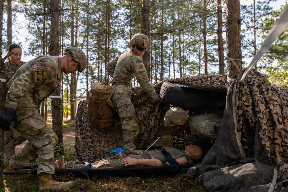 Military personnel in MultiCam/OCP camouflage uniforms are conducting a combat casualty care training exercise in a forested area. A simulated casualty is lying on a litter/stretcher while multiple soldiers attend to them near a camouflage net shelter with a tire prop. The scene appears to be a field medical training scenario, likely a tactical combat casualty care (TCCC) exercise.