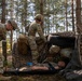 Military personnel in MultiCam/OCP camouflage uniforms are conducting a combat casualty care training exercise in a forested area. A simulated casualty is lying on a litter/stretcher while multiple soldiers attend to them near a camouflage net shelter with a tire prop. The scene appears to be a field medical training scenario, likely a tactical combat casualty care (TCCC) exercise.
