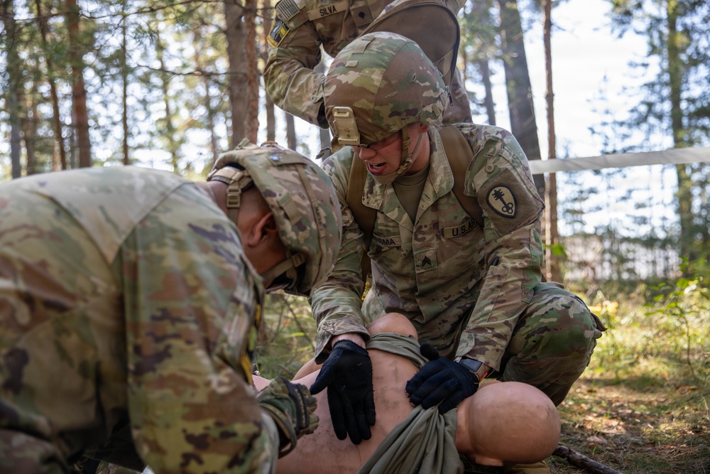 U.S. Army soldiers in MultiCam/OCP camouflage uniforms are conducting a tactical combat casualty care (TCCC) training exercise in a forested environment. At least two soldiers are actively treating a simulated casualty (mannequin or training dummy) on the ground, applying what appears to be a tourniquet or bandage to the lower extremities. A third soldier identified by the name tape 'SILVA' is visible in the background providing oversight or assistance.
