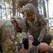U.S. Army soldiers in MultiCam/OCP camouflage uniforms are conducting a tactical combat casualty care (TCCC) training exercise in a forested environment. At least two soldiers are actively treating a simulated casualty (mannequin or training dummy) on the ground, applying what appears to be a tourniquet or bandage to the lower extremities. A third soldier identified by the name tape 'SILVA' is visible in the background providing oversight or assistance.