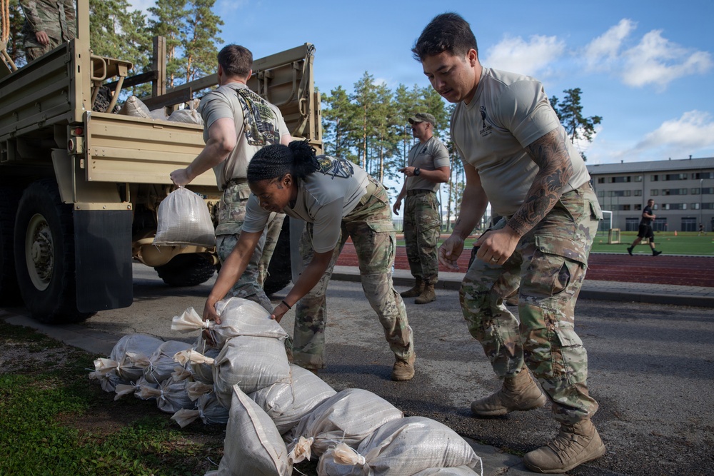 Military personnel in U.S. Army OCP (Operational Camouflage Pattern) uniforms are unloading and stacking sandbags from a military cargo truck. The activity appears to be flood control or base preparation work, taking place on or near a running track at what appears to be a military installation. Multiple soldiers are working cooperatively, with some on the truck bed and others stacking bags on the ground.