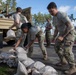 Military personnel in U.S. Army OCP (Operational Camouflage Pattern) uniforms are unloading and stacking sandbags from a military cargo truck. The activity appears to be flood control or base preparation work, taking place on or near a running track at what appears to be a military installation. Multiple soldiers are working cooperatively, with some on the truck bed and others stacking bags on the ground.