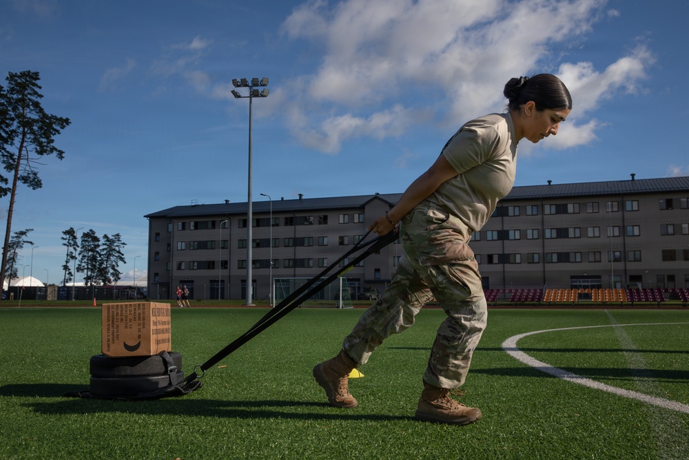 A female soldier in military camouflage uniform is performing a physical training exercise on an artificial turf sports field, dragging a weighted sled loaded with tires and a cardboard box using resistance straps. The setting appears to be a military installation or base, evidenced by the institutional buildings and sports facilities visible in the background. The exercise is a common military fitness drill designed to build strength and endurance.