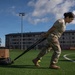 A female soldier in military camouflage uniform is performing a physical training exercise on an artificial turf sports field, dragging a weighted sled loaded with tires and a cardboard box using resistance straps. The setting appears to be a military installation or base, evidenced by the institutional buildings and sports facilities visible in the background. The exercise is a common military fitness drill designed to build strength and endurance.