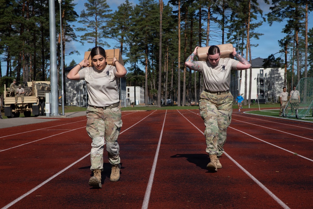 Two female military personnel in camouflage uniforms are carrying weighted sandbags or ruck loads on their shoulders while walking on a red athletic track. They appear to be participating in a physical fitness or military training event, likely a Wounded Warrior or Army fitness competition based on the visible logo on their shirts. The setting appears to be a military installation with tall pine trees, a white building, and military vehicles visible in the background.