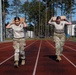 Two female military personnel in camouflage uniforms are carrying weighted sandbags or ruck loads on their shoulders while walking on a red athletic track. They appear to be participating in a physical fitness or military training event, likely a Wounded Warrior or Army fitness competition based on the visible logo on their shirts. The setting appears to be a military installation with tall pine trees, a white building, and military vehicles visible in the background.