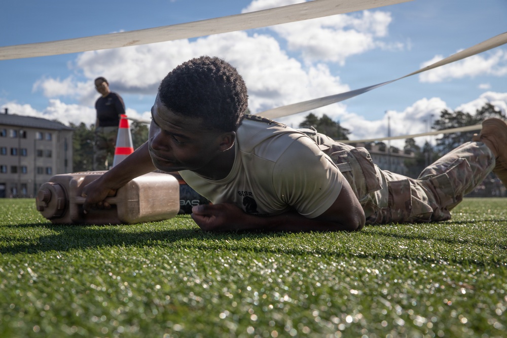 A soldier in military uniform is crawling under a low wire or rope obstacle on an artificial turf field, pushing a weighted object (dumbbell or log) in front of them. This appears to be a military fitness or obstacle course event, likely a competition or training exercise. A second person in dark clothing stands in the background near an orange traffic cone.
