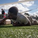 A soldier in military uniform is crawling under a low wire or rope obstacle on an artificial turf field, pushing a weighted object (dumbbell or log) in front of them. This appears to be a military fitness or obstacle course event, likely a competition or training exercise. A second person in dark clothing stands in the background near an orange traffic cone.