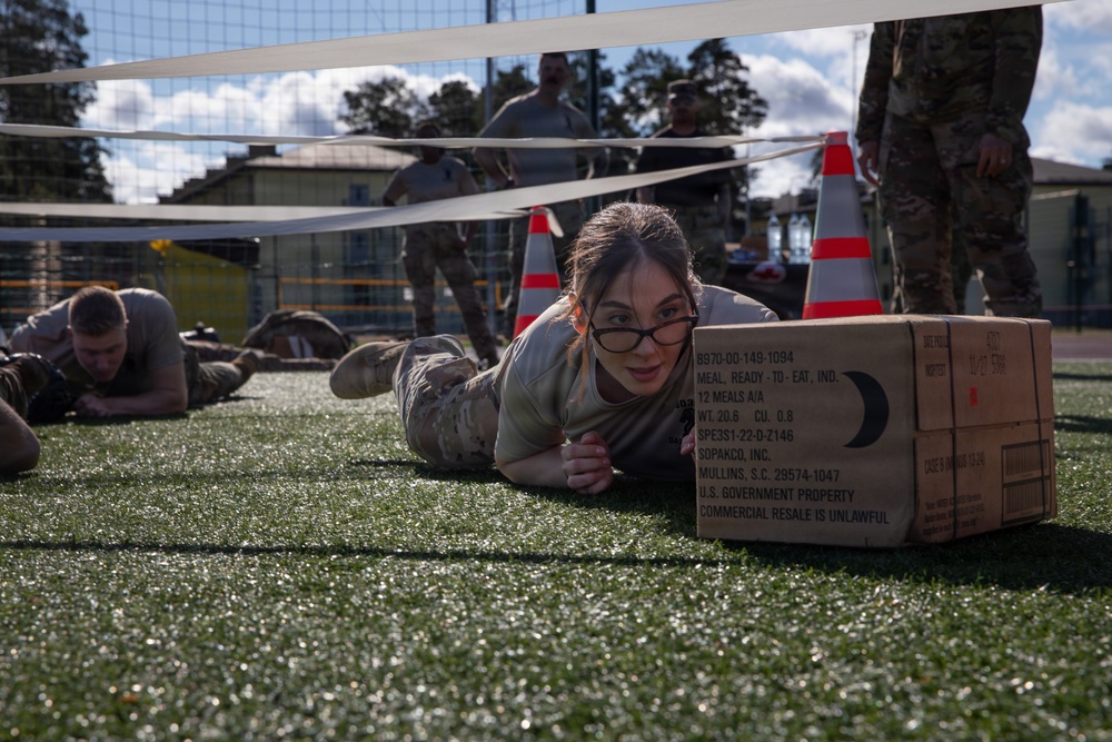 A female soldier in military camouflage uniform is crawling under a low net on an artificial turf field while pushing a cardboard MRE (Meal Ready to Eat) box. This appears to be a military obstacle course or physical training exercise, with other soldiers visible crawling in the background and observers standing nearby. Orange traffic cones mark the course boundaries.