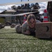 A female soldier in military camouflage uniform is crawling under a low net on an artificial turf field while pushing a cardboard MRE (Meal Ready to Eat) box. This appears to be a military obstacle course or physical training exercise, with other soldiers visible crawling in the background and observers standing nearby. Orange traffic cones mark the course boundaries.