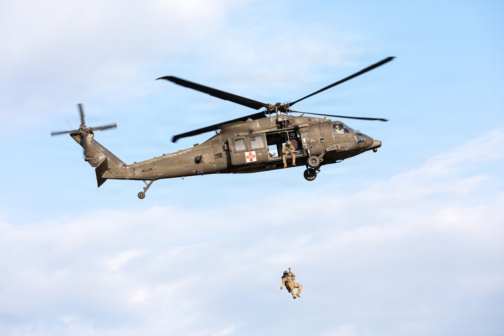 A military UH-60 Black Hawk helicopter marked with a red cross medical insignia is airborne against a partly cloudy sky. A soldier or crew member is being lowered or hoisted on a cable beneath the aircraft, while another personnel is visible seated at the open side door. This appears to be a medical evacuation (MEDEVAC) training exercise or hoist rescue operation.