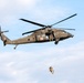 A military UH-60 Black Hawk helicopter marked with a red cross medical insignia is airborne against a partly cloudy sky. A soldier or crew member is being lowered or hoisted on a cable beneath the aircraft, while another personnel is visible seated at the open side door. This appears to be a medical evacuation (MEDEVAC) training exercise or hoist rescue operation.