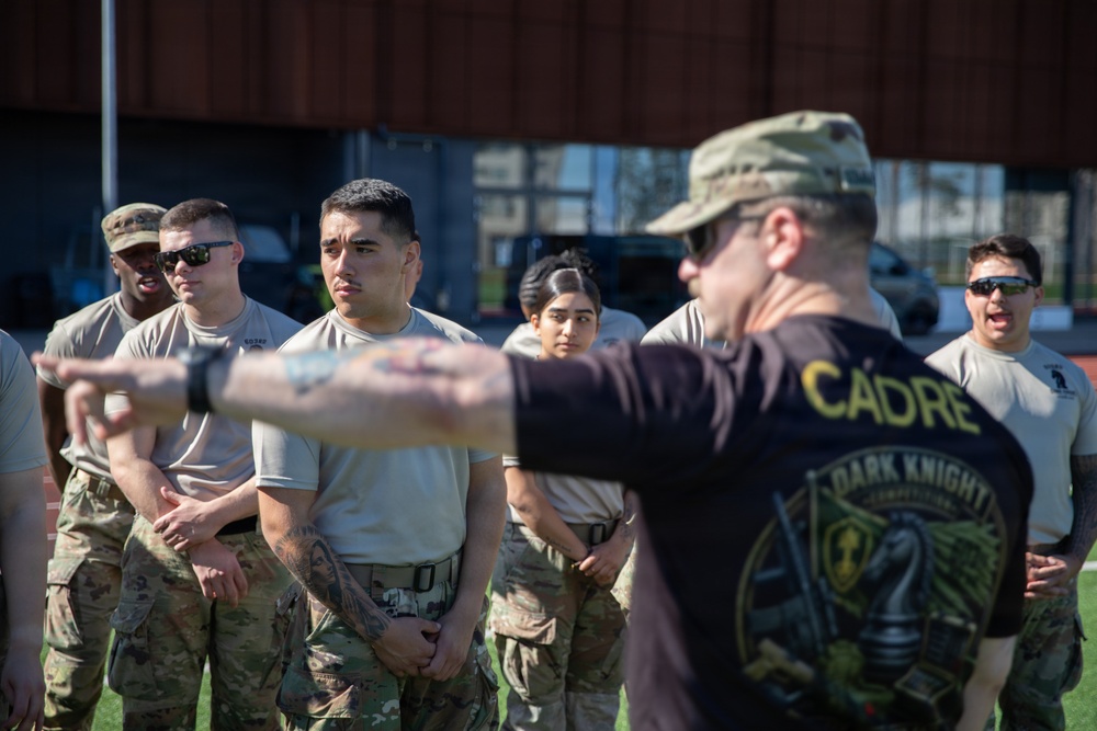 A military cadre instructor is briefing or directing a group of soldiers during what appears to be a training exercise or competition. The instructor, wearing a black 'CADRE' shirt with 'Dark Knight Competition' branding, is pointing and giving instructions to uniformed soldiers. The soldiers are wearing OCP (Operational Camouflage Pattern) uniforms and appear attentive.
