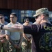 A military cadre instructor is briefing or directing a group of soldiers during what appears to be a training exercise or competition. The instructor, wearing a black 'CADRE' shirt with 'Dark Knight Competition' branding, is pointing and giving instructions to uniformed soldiers. The soldiers are wearing OCP (Operational Camouflage Pattern) uniforms and appear attentive.