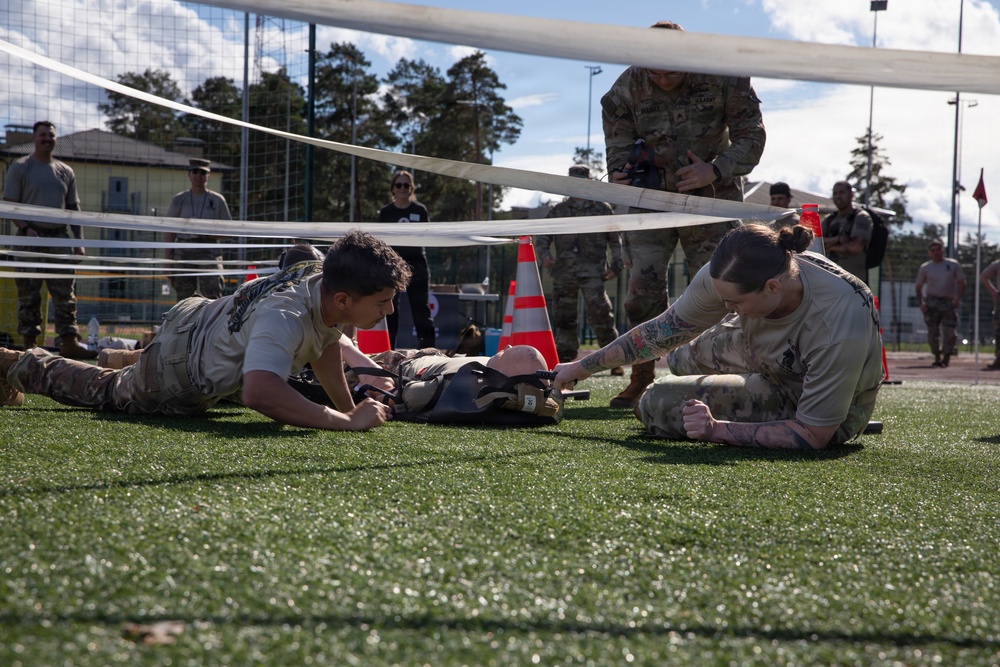 Two military personnel in camouflage uniforms are performing what appears to be a combat casualty care or tactical combat training exercise on an artificial turf field. They are attending to a simulated casualty lying on the ground beneath a net, with one soldier on the left and another with visible tattoos on the right. Observers and additional military personnel are visible in the background, suggesting this is a supervised training event or competition.