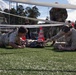 Two military personnel in camouflage uniforms are performing what appears to be a combat casualty care or tactical combat training exercise on an artificial turf field. They are attending to a simulated casualty lying on the ground beneath a net, with one soldier on the left and another with visible tattoos on the right. Observers and additional military personnel are visible in the background, suggesting this is a supervised training event or competition.