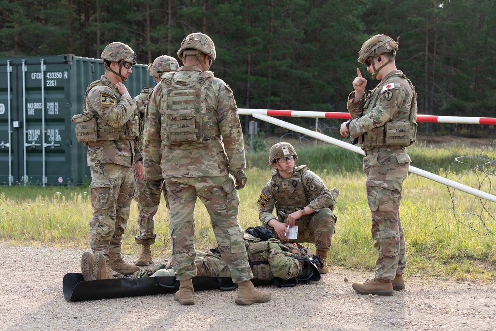 A group of military personnel in camouflage uniforms are gathered at what appears to be a checkpoint or training area near a barrier gate. One soldier wearing a red cross medical insignia is gesturing with one finger while another soldier kneels on the ground attending to a simulated casualty or equipment on a litter. The scene appears to be a medical training exercise at a military installation.