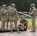 A group of military personnel in camouflage uniforms are gathered at what appears to be a checkpoint or training area near a barrier gate. One soldier wearing a red cross medical insignia is gesturing with one finger while another soldier kneels on the ground attending to a simulated casualty or equipment on a litter. The scene appears to be a medical training exercise at a military installation.