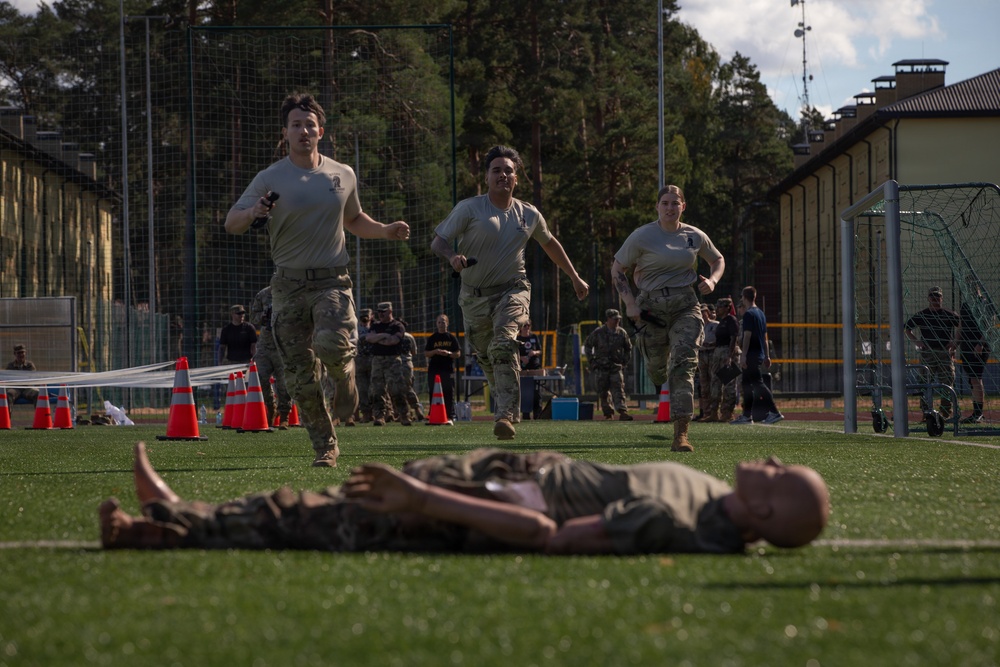 Military personnel in U.S. Army combat uniforms (OCP pattern) are participating in what appears to be a physical fitness competition or training event on an artificial turf field. Three soldiers are running toward the camera while a fourth soldier lies flat on their back on the ground in the foreground, likely having just completed a physical exercise. Orange traffic cones mark a course boundary, and numerous spectators and other military personnel are visible in the background.