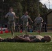 Military personnel in U.S. Army combat uniforms (OCP pattern) are participating in what appears to be a physical fitness competition or training event on an artificial turf field. Three soldiers are running toward the camera while a fourth soldier lies flat on their back on the ground in the foreground, likely having just completed a physical exercise. Orange traffic cones mark a course boundary, and numerous spectators and other military personnel are visible in the background.