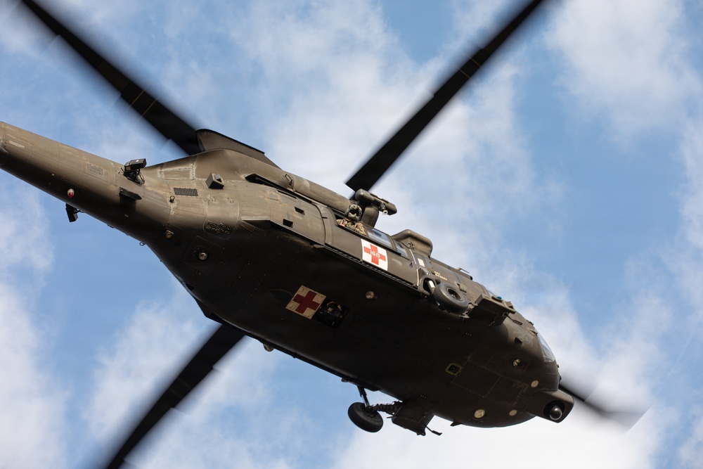 A military medical evacuation (MEDEVAC) helicopter is photographed from directly below as it flies overhead. The aircraft displays red cross markings on its fuselage, identifying it as a medical/rescue helicopter. The image is taken from a ground-up perspective, capturing the underside of the aircraft against a partly cloudy blue sky.