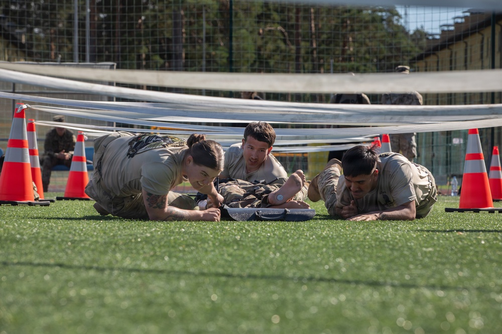 Three military personnel in olive/tan uniforms are crawling under a low barrier on an artificial turf field, performing what appears to be a low-crawl obstacle course exercise. Orange traffic cones mark the boundaries of the obstacle lane. The setting appears to be a military training or competition event at an outdoor sports facility.
