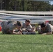 Three military personnel in olive/tan uniforms are crawling under a low barrier on an artificial turf field, performing what appears to be a low-crawl obstacle course exercise. Orange traffic cones mark the boundaries of the obstacle lane. The setting appears to be a military training or competition event at an outdoor sports facility.