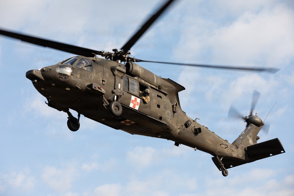 A military medical evacuation (MEDEVAC) helicopter, identifiable as a Sikorsky UH-60 Black Hawk, is captured in flight against a partly cloudy blue sky. The aircraft is painted in olive drab/dark green military livery and displays a prominent red cross emblem on its fuselage, indicating its medical role. A crew member is visible in the open side door of the helicopter.