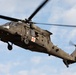A military medical evacuation (MEDEVAC) helicopter, identifiable as a Sikorsky UH-60 Black Hawk, is captured in flight against a partly cloudy blue sky. The aircraft is painted in olive drab/dark green military livery and displays a prominent red cross emblem on its fuselage, indicating its medical role. A crew member is visible in the open side door of the helicopter.