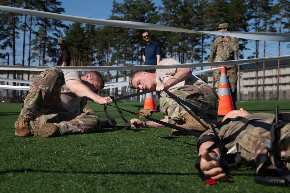 Military personnel in camouflage uniforms are participating in what appears to be a physical training or obstacle course exercise on an artificial turf field. Two soldiers in the foreground are crawling under a low horizontal bar or wire obstacle while pulling straps attached to a third soldier lying on the ground. Observers in civilian and military attire watch from the background near orange traffic cones and a fenced perimeter.