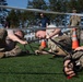 Military personnel in camouflage uniforms are participating in what appears to be a physical training or obstacle course exercise on an artificial turf field. Two soldiers in the foreground are crawling under a low horizontal bar or wire obstacle while pulling straps attached to a third soldier lying on the ground. Observers in civilian and military attire watch from the background near orange traffic cones and a fenced perimeter.