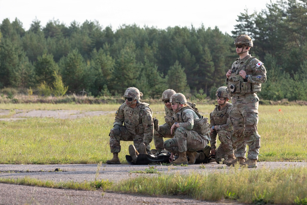 A group of military personnel in camouflage uniforms are conducting what appears to be a tactical combat casualty care (TCCC) training exercise in an open field area. Four soldiers are attending to a simulated casualty on the ground while a fifth soldier, identifiable by a red cross medical patch, stands observing the scenario. The setting appears to be a military training area with a paved surface, open grassland, and a tree line in the background.