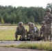 A group of military personnel in camouflage uniforms are conducting what appears to be a tactical combat casualty care (TCCC) training exercise in an open field area. Four soldiers are attending to a simulated casualty on the ground while a fifth soldier, identifiable by a red cross medical patch, stands observing the scenario. The setting appears to be a military training area with a paved surface, open grassland, and a tree line in the background.