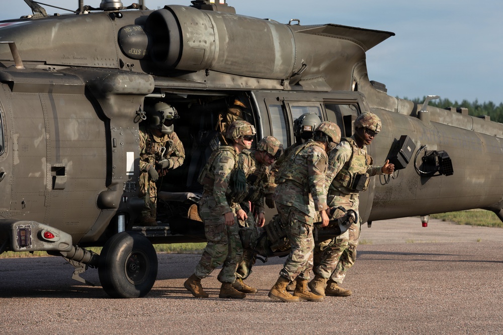 Military personnel in camouflage uniforms are conducting what appears to be a MEDEVAC or casualty evacuation exercise beside a UH-60 Black Hawk helicopter on a tarmac. Several soldiers are carrying or supporting what appears to be a casualty on a litter toward the open side door of the aircraft. A crew member remains inside the helicopter while ground personnel assist with the loading procedure.