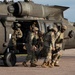Military personnel in camouflage uniforms are conducting what appears to be a MEDEVAC or casualty evacuation exercise beside a UH-60 Black Hawk helicopter on a tarmac. Several soldiers are carrying or supporting what appears to be a casualty on a litter toward the open side door of the aircraft. A crew member remains inside the helicopter while ground personnel assist with the loading procedure.