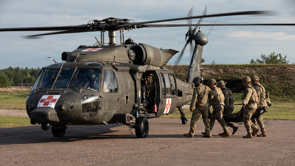 A military medical evacuation (MEDEVAC) operation is depicted, showing soldiers carrying a casualty on a litter toward a UH-60 Black Hawk helicopter marked with red cross insignia. The helicopter is running with rotors spinning, indicating an active or imminent extraction. The scene appears to be a training exercise or real MEDEVAC operation conducted on a paved landing area.