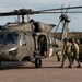 A military medical evacuation (MEDEVAC) operation is depicted, showing soldiers carrying a casualty on a litter toward a UH-60 Black Hawk helicopter marked with red cross insignia. The helicopter is running with rotors spinning, indicating an active or imminent extraction. The scene appears to be a training exercise or real MEDEVAC operation conducted on a paved landing area.
