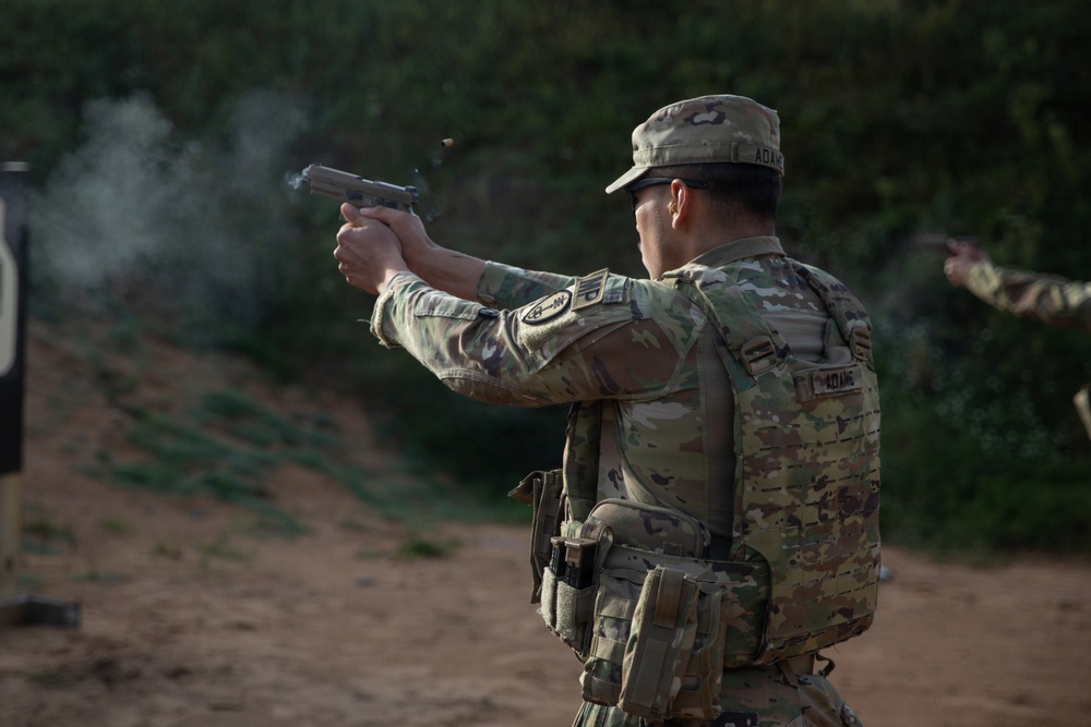 A military or law enforcement personnel wearing OCP (Operational Camouflage Pattern) uniform and body armor is actively firing a semi-automatic pistol at a shooting range. A spent brass casing is visible ejecting from the firearm, and smoke is present near the muzzle. The name tape on the uniform reads 'ADAMS'.