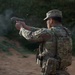 A military or law enforcement personnel wearing OCP (Operational Camouflage Pattern) uniform and body armor is actively firing a semi-automatic pistol at a shooting range. A spent brass casing is visible ejecting from the firearm, and smoke is present near the muzzle. The name tape on the uniform reads 'ADAMS'.