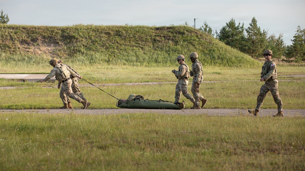 Four military personnel in camouflage uniforms are conducting what appears to be a casualty evacuation (CASEVAC) drill on a training ground. Two soldiers are dragging a green military litter/stretcher along a paved path while two others walk alongside. The setting appears to be a military training area with earthen berms and tree lines in the background.