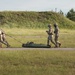 Four military personnel in camouflage uniforms are conducting what appears to be a casualty evacuation (CASEVAC) drill on a training ground. Two soldiers are dragging a green military litter/stretcher along a paved path while two others walk alongside. The setting appears to be a military training area with earthen berms and tree lines in the background.