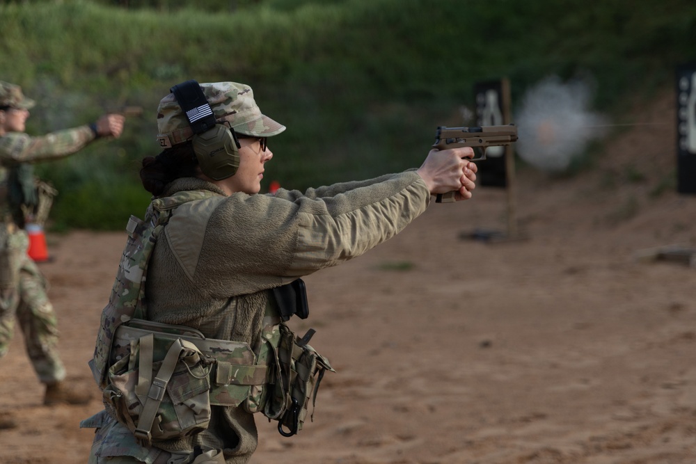 A military or law enforcement personnel is actively firing a pistol at a shooting range, with visible muzzle blast at the target downrange. The shooter is wearing full tactical gear including a multicam uniform, plate carrier, ear protection, and eye protection with a US flag patch visible on the cap. Additional personnel in similar uniforms are visible in the background, suggesting a group training exercise.