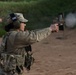 A military or law enforcement personnel is actively firing a pistol at a shooting range, with visible muzzle blast at the target downrange. The shooter is wearing full tactical gear including a multicam uniform, plate carrier, ear protection, and eye protection with a US flag patch visible on the cap. Additional personnel in similar uniforms are visible in the background, suggesting a group training exercise.