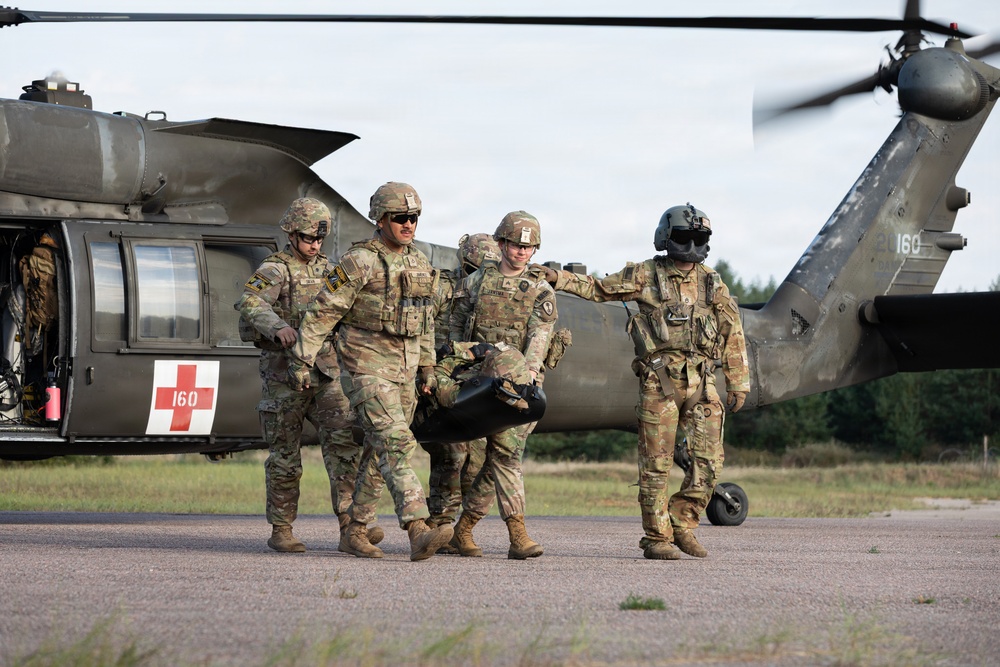 Military personnel in camouflage uniforms are carrying a casualty or equipment bag away from a medical evacuation (MEDEVAC) helicopter marked with a red cross and the number '160'. The helicopter appears to be a UH-60 Black Hawk, identifiable by its distinctive silhouette and olive drab paint scheme. The scene appears to depict a MEDEVAC training exercise or operational casualty evacuation on a tarmac or landing pad.