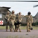Military personnel in camouflage uniforms are carrying a casualty or equipment bag away from a medical evacuation (MEDEVAC) helicopter marked with a red cross and the number '160'. The helicopter appears to be a UH-60 Black Hawk, identifiable by its distinctive silhouette and olive drab paint scheme. The scene appears to depict a MEDEVAC training exercise or operational casualty evacuation on a tarmac or landing pad.
