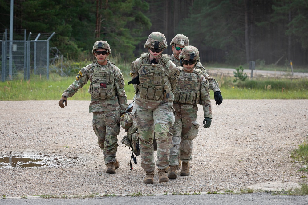 Four military personnel in U.S. Army OCP (Operational Camouflage Pattern) uniforms are walking along a gravel path in what appears to be a training area or military installation. Two soldiers in the center appear to be assisting or supporting a third soldier, possibly simulating a casualty evacuation drill. The setting includes a forested background, a perimeter fence, and a gravel road.