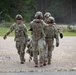 Four military personnel in U.S. Army OCP (Operational Camouflage Pattern) uniforms are walking along a gravel path in what appears to be a training area or military installation. Two soldiers in the center appear to be assisting or supporting a third soldier, possibly simulating a casualty evacuation drill. The setting includes a forested background, a perimeter fence, and a gravel road.