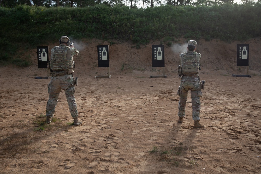 Two military personnel in full combat gear and camouflage uniforms are conducting pistol qualification or training at an outdoor shooting range. The left shooter is actively firing, as evidenced by visible muzzle smoke, while the right shooter stands in a ready position. Four standard silhouette targets are positioned downrange against a sandy berm backdrop.
